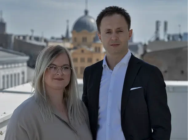 Piotr Nowakowski i Dominika Sujka-Kujawiak elegancko ubrani, na tle nowoczesnych budynków. Jeden z nich ma kopułę i ozdobne kolumny. Oboje patrzą się poważnie w obiektyw