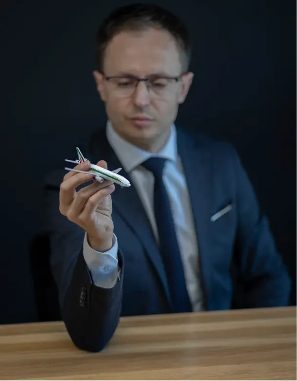 Piotr Nowakowski w eleganckim, granatowym garniturze. Siedzi on przy drewnianym biurku i trzyma w ręce małą figurkę samolotu. Zdjęcie to nawiązuje do hałasu lotniczego.