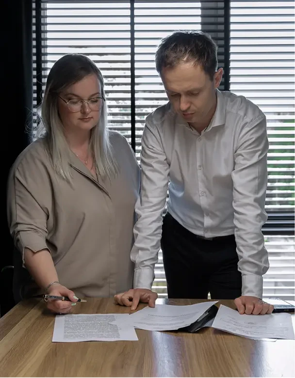 Elegancko ubrani adwokaci z Kancelarii Nowakowski – Piotr i Dominika, którzy przeglądają ważne dokumenty w biurze przy drewnianym biurku.