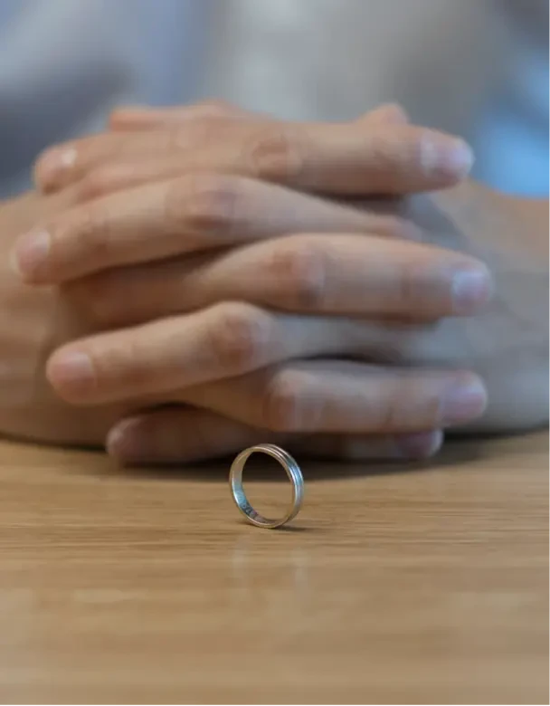 Splecione dłonie osoby, która siedzi przy biurku, na którym leży obrączka. Całość symbolizuje sprawy rozwodowe.