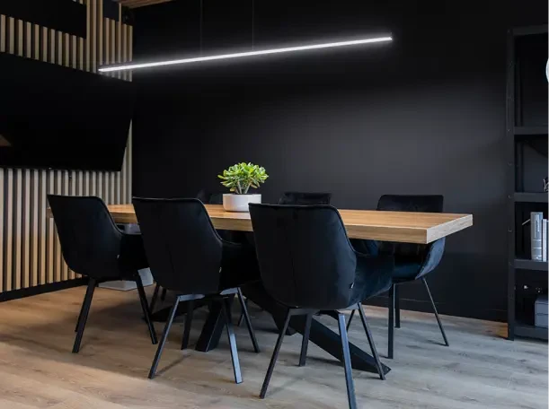 Wnętrze kancelarii adwokackiej Nowakowski&Sujka-Kujawiak. Nowoczesne wnętrze, solidny, drewniany stół z sześcioma czarnymi krzesłami przystawionymi do niego. Na podłodze nowoczesne szare panele, ściany są pomalowane na czarno, ale pomieszczenie jest przyjemnie oświetlone i tworzy atmosferę skupienia, komfortu i luksusu. Jest to przestronne pomieszczenie z pięknym wystrojem